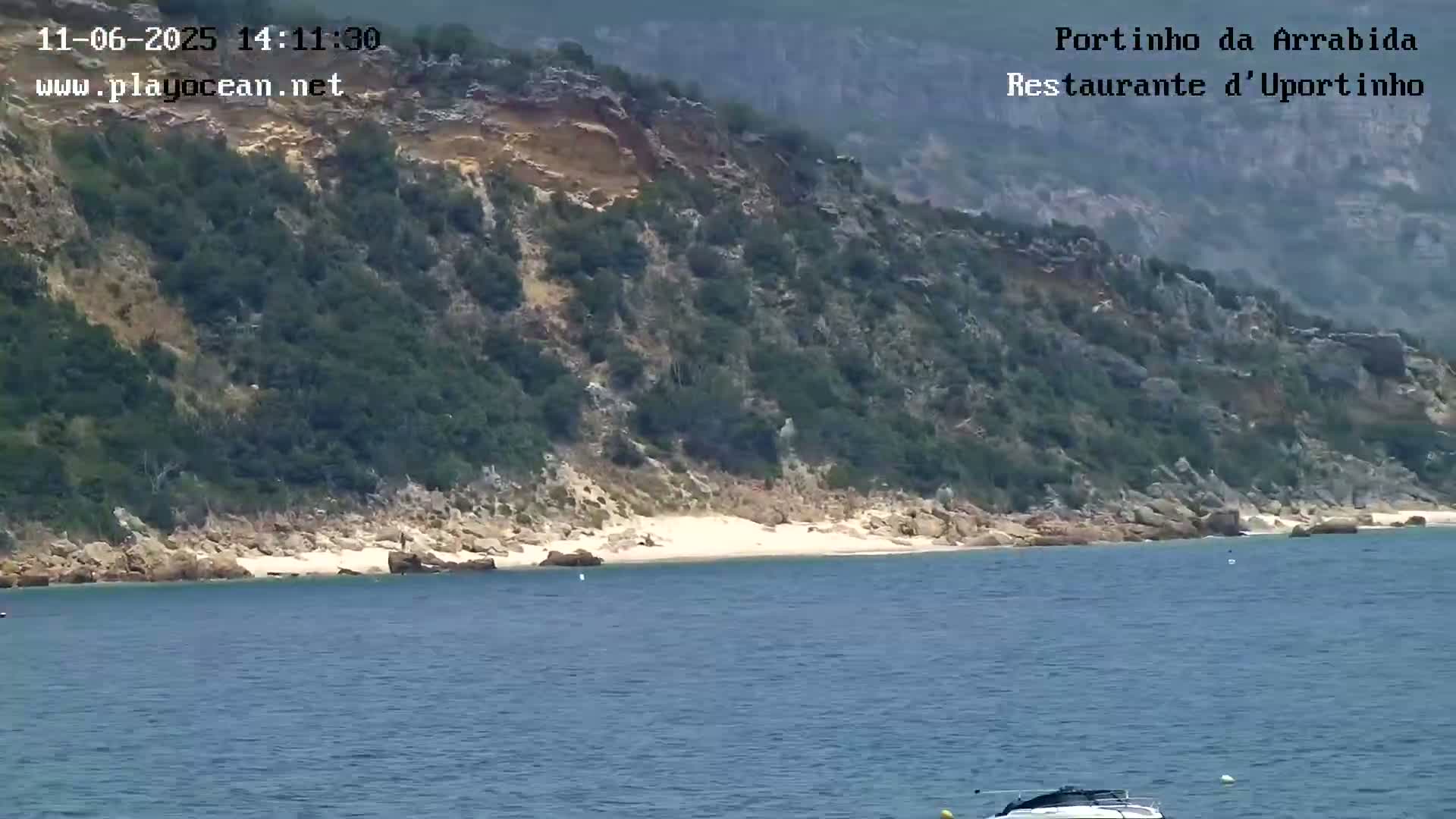 A small, sandy cove is nestled below a rocky, green hillside overlooking calm blue water with a boat visible in the distance.