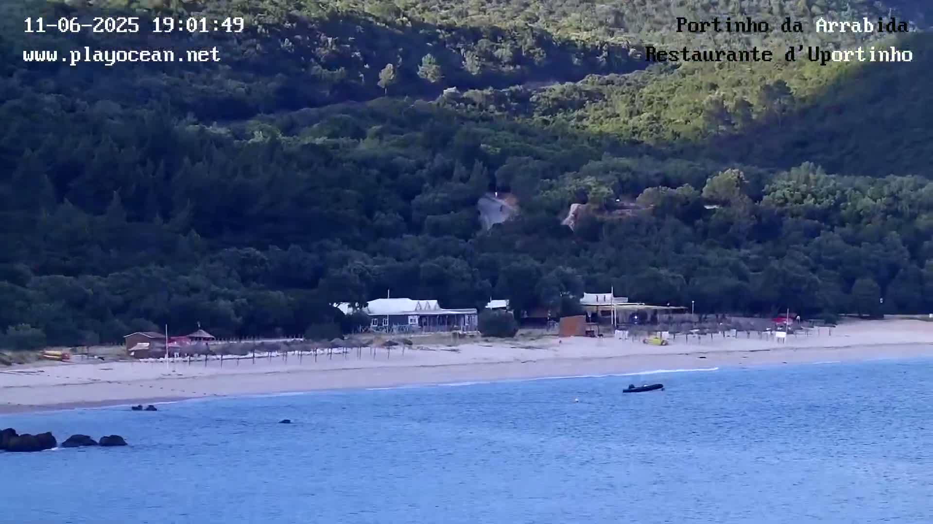 A sandy beach with a few buildings and a small boat in calm water, backed by a lush, green hillside under a clear sky.