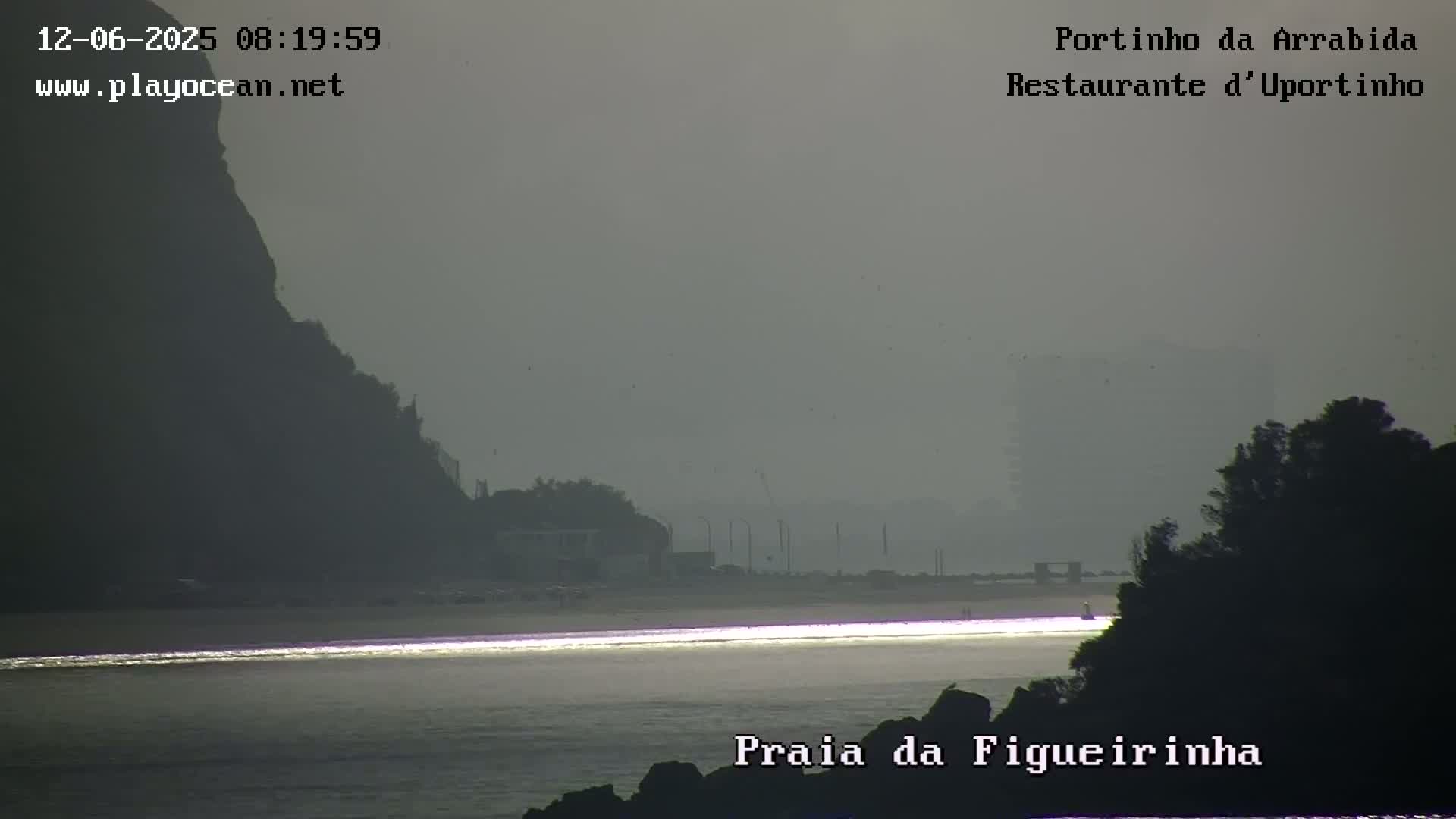 A hazy, overcast day shows a calm body of water reflecting light near a beach with a dark hill or cliff in the background.
