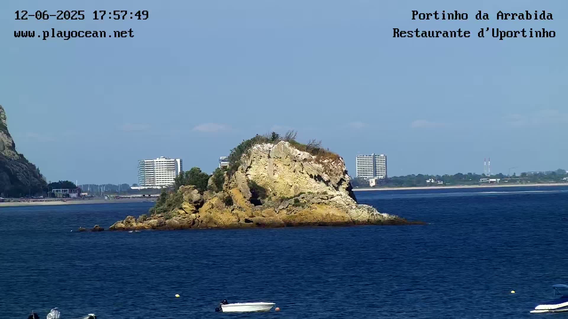 A rocky islet sits in calm blue water near a beach and distant buildings under a clear blue sky.