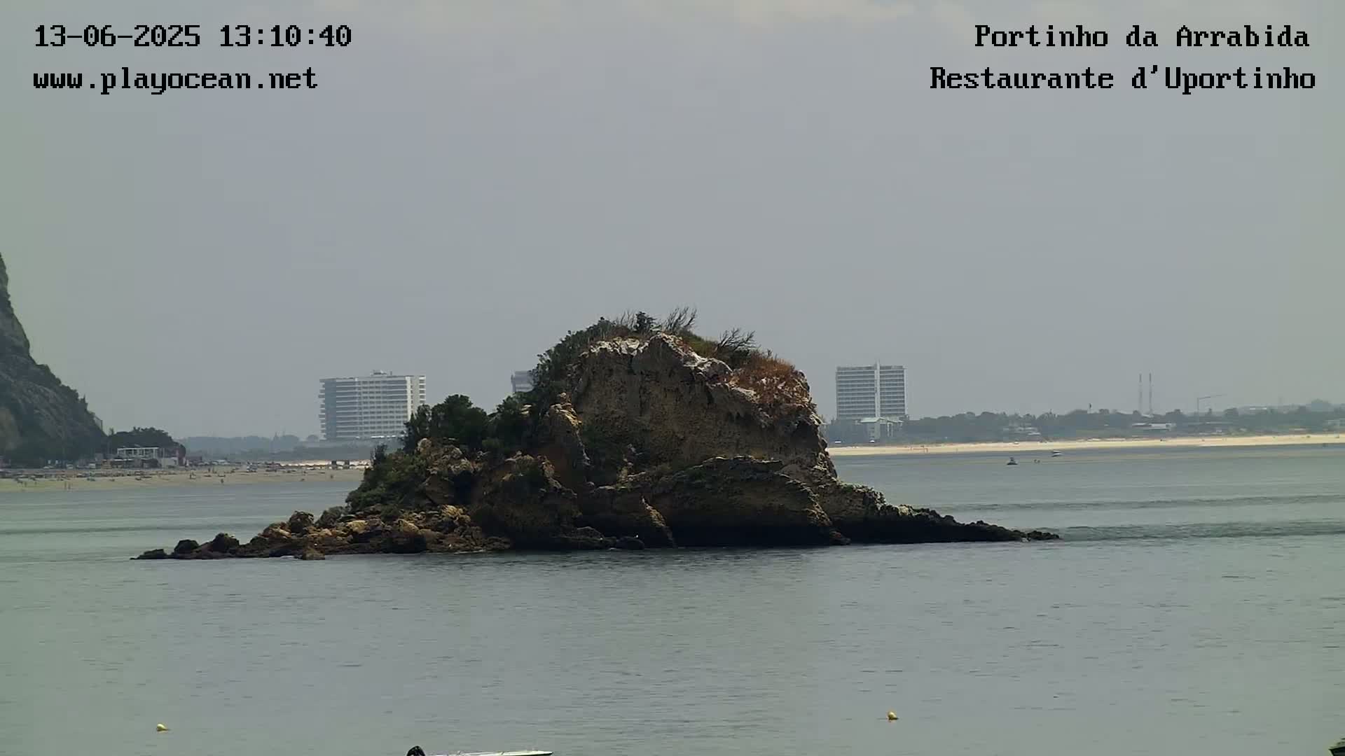 A rocky outcrop sits in calm, gray water near a sandy beach and distant buildings under an overcast sky.