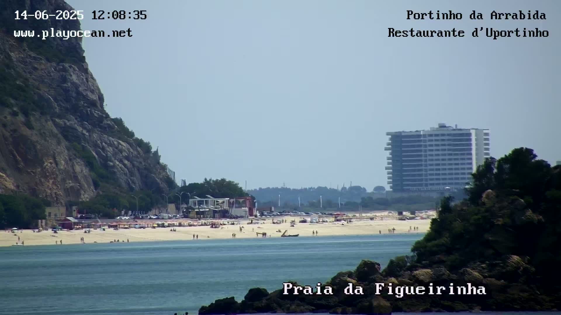A sandy beach with people on it is visible, along with a large building in the background, a rocky cliff to the left, and calm water in the foreground under a clear sky.
