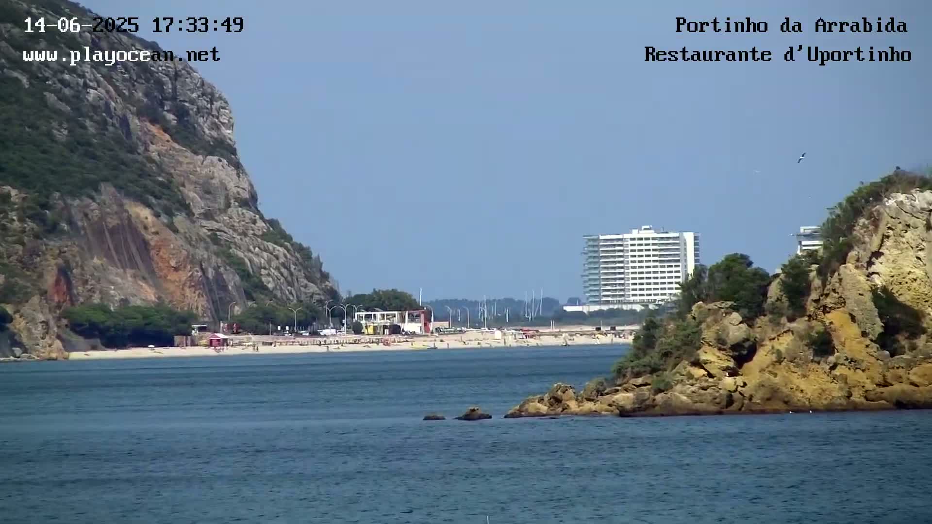 A sandy beach with people on it sits between rocky hills, one of which is partially covered with vegetation, under a clear blue sky.