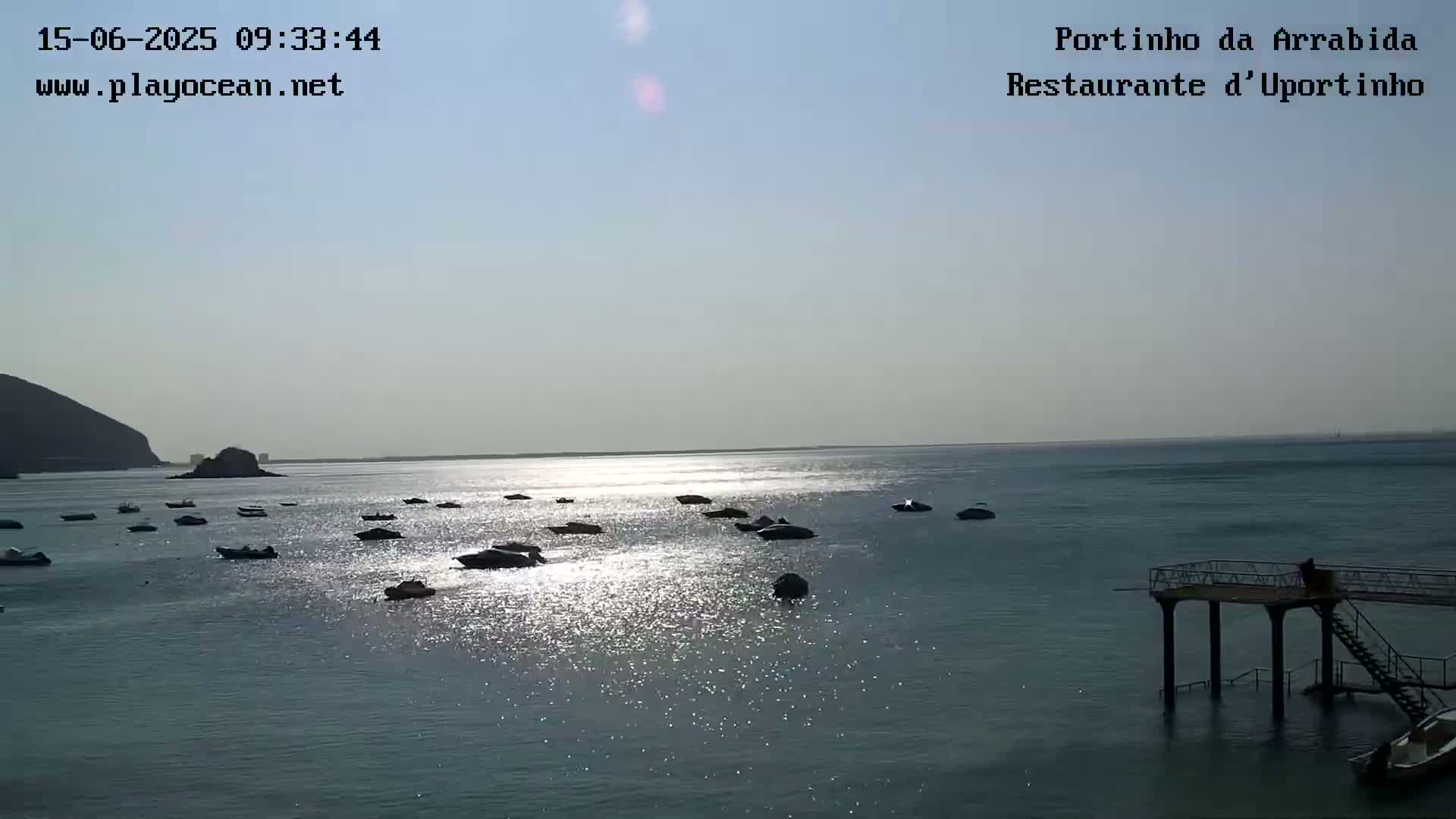 A calm, sunny day shows numerous small boats moored in a bay near a rocky shore and a small pier.