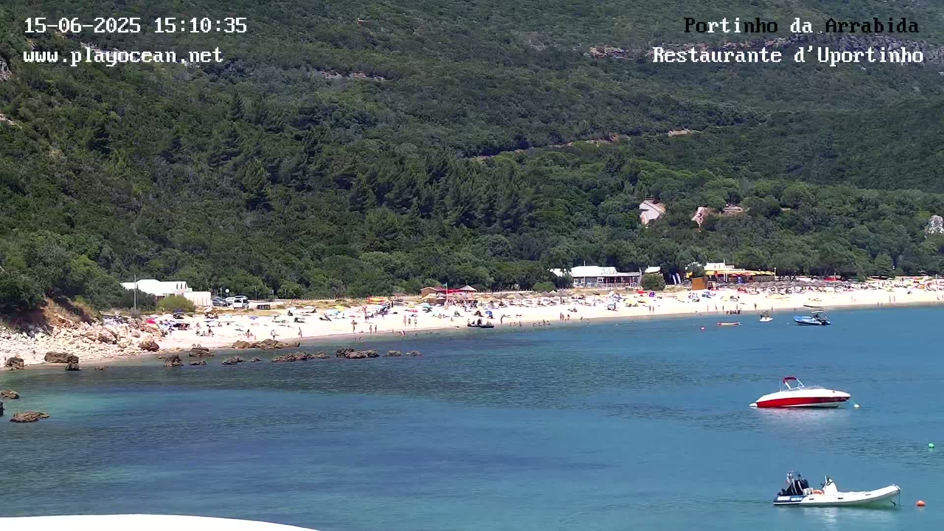 A sandy beach with many people on it is nestled in a cove bordered by lush green hills, with several small boats in the calm, clear water.