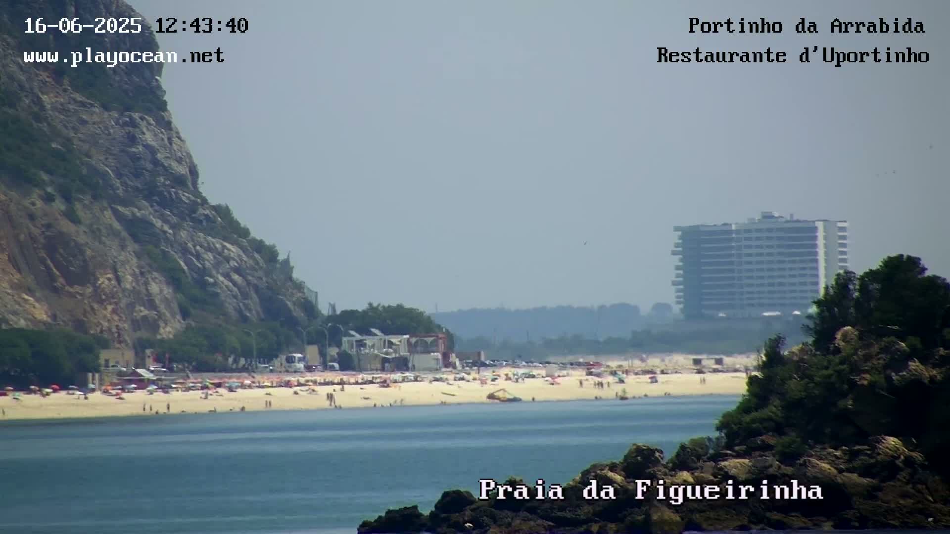 A hazy, sunny day shows a crowded beach with a rocky cliff to the left and a tall building in the distance.