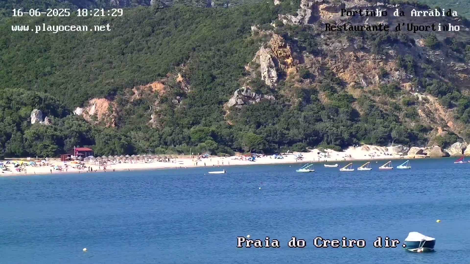 A sandy beach with many people and several small boats in calm blue water, backed by green hills under a sunny sky.