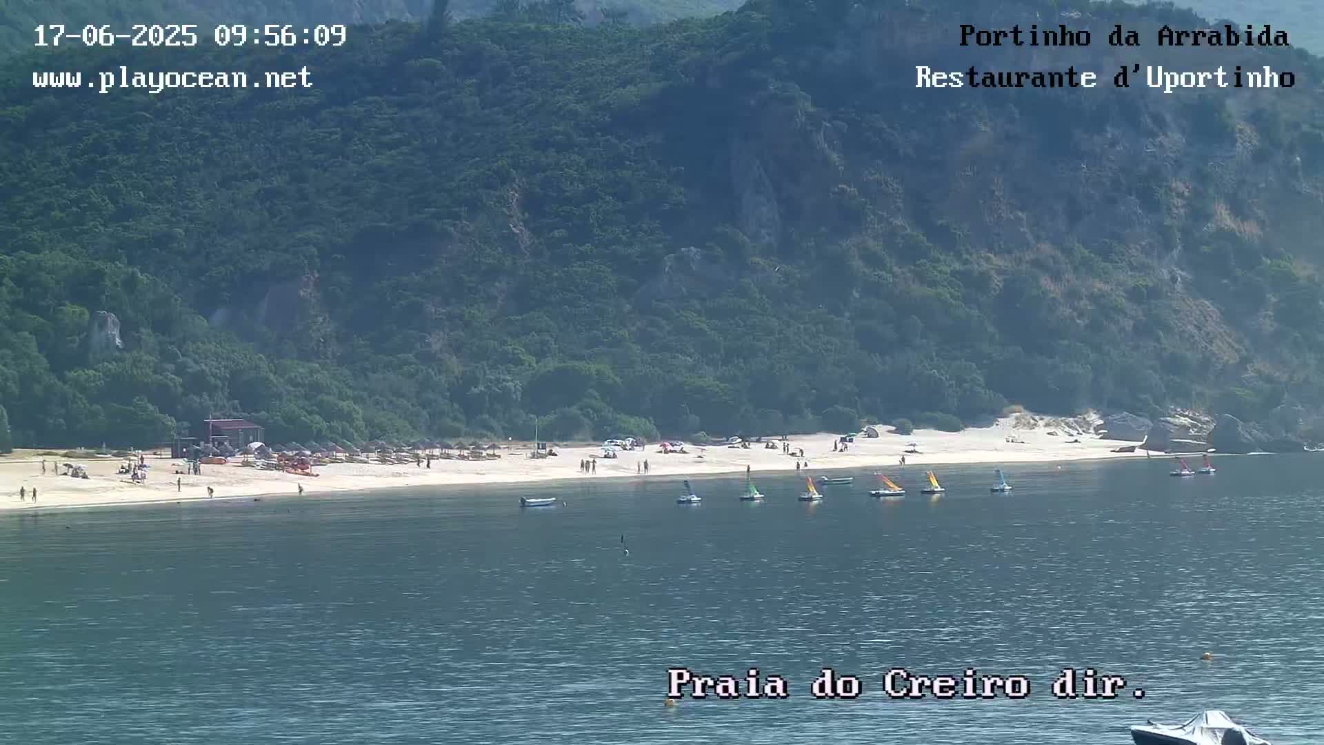 A sandy beach with a few people and several small sailboats in calm water, backed by lush green hills under a clear sky.