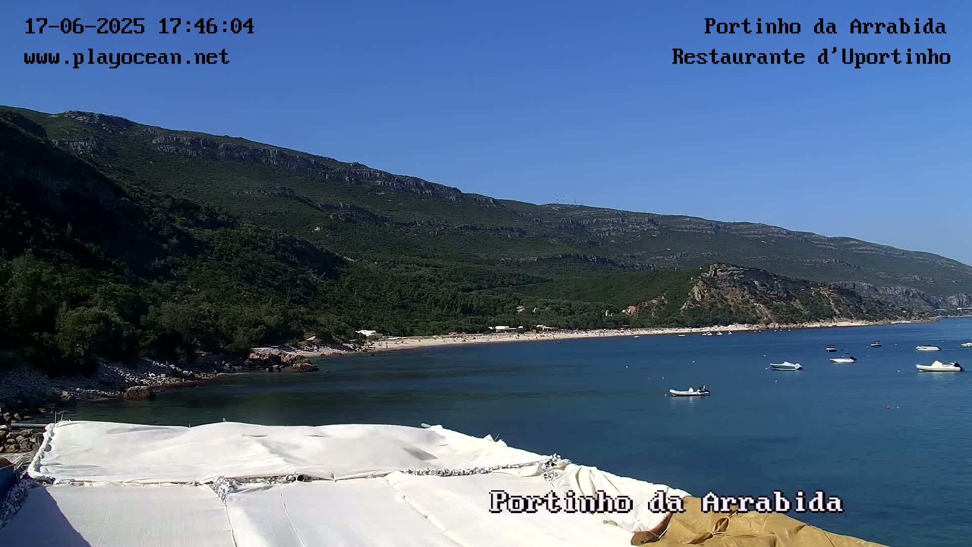 A sunny day overlooks a calm bay with several small boats, a sandy beach lined with people, and a mountainous, green landscape in the background.