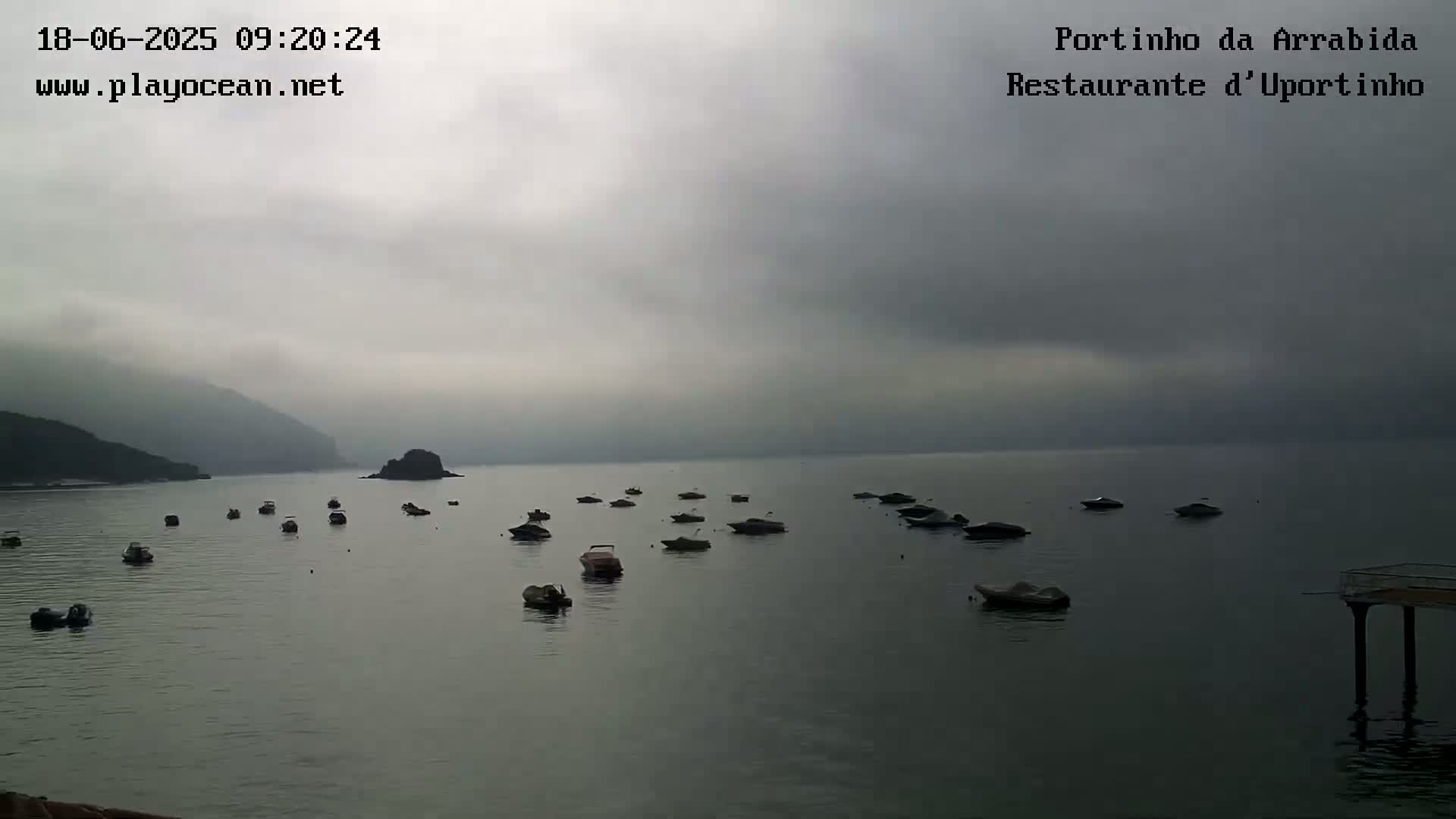 Many small boats are scattered across a calm bay on an overcast day, with hills shrouded in fog in the background.