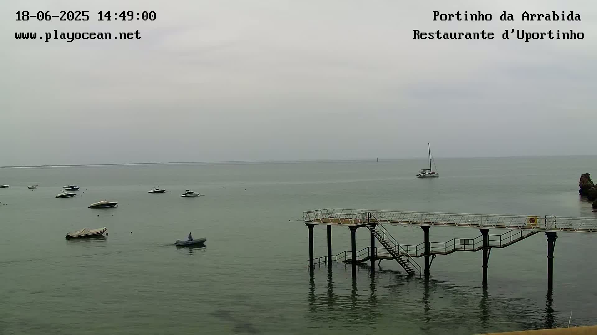 A calm, overcast day shows several small boats moored in a bay, with a larger sailboat further out, and a pier extending into the water.