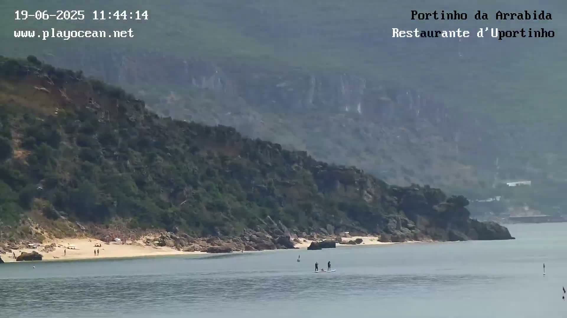 A hazy, sunny day shows a calm bay with a sandy beach, green hills, and two people paddleboarding.