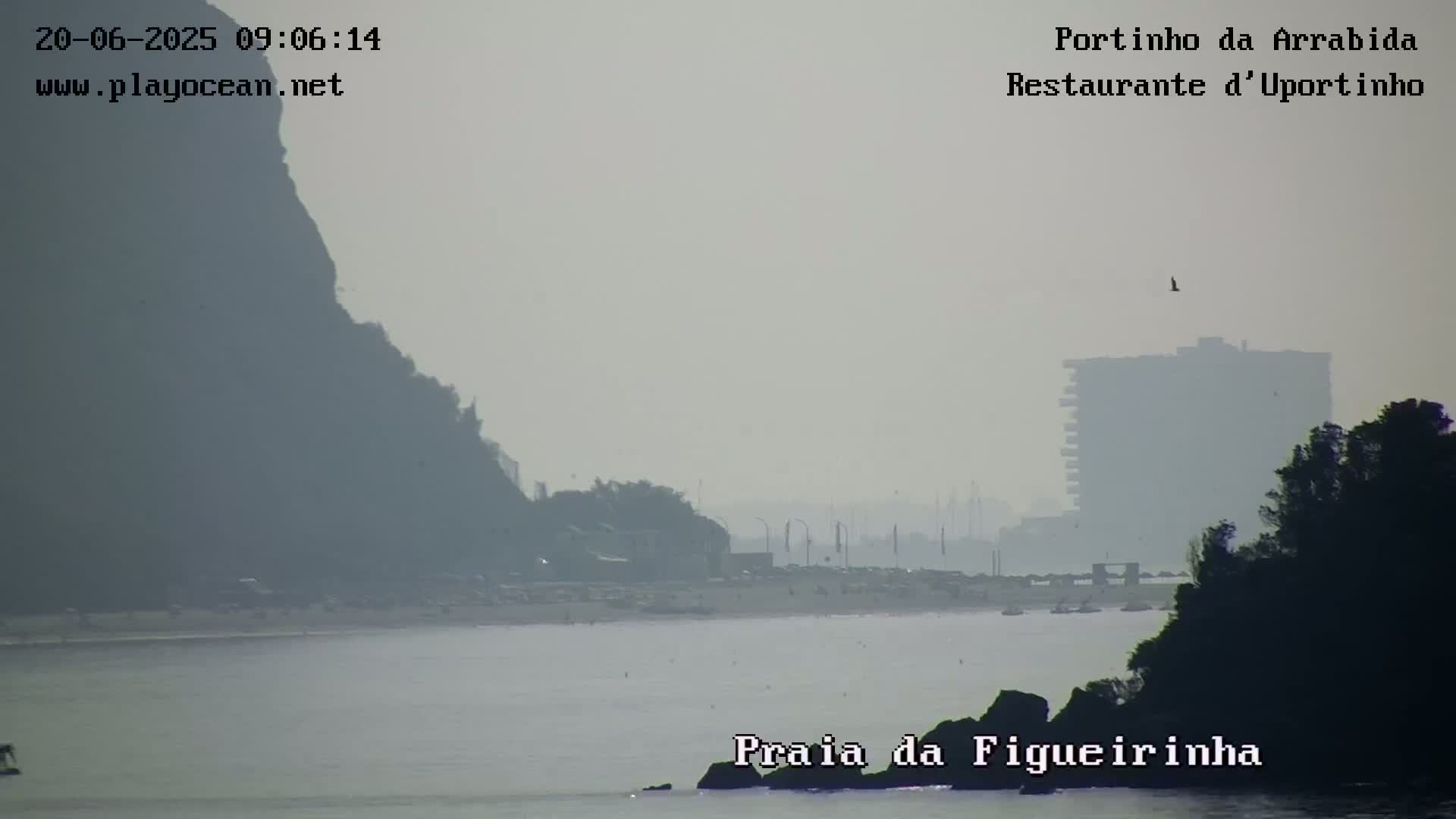 A hazy, overcast view shows a beach with dark hills and a building in the distance.