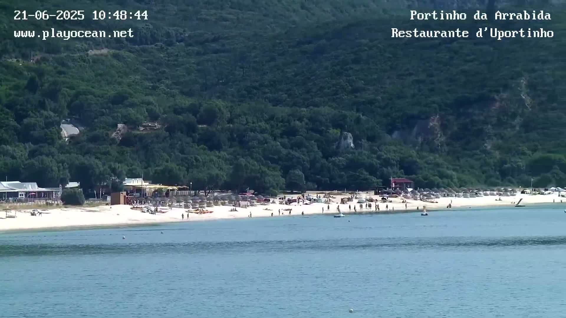 A sandy beach with people and beach umbrellas is nestled between calm water and a lush green hillside on a sunny day.