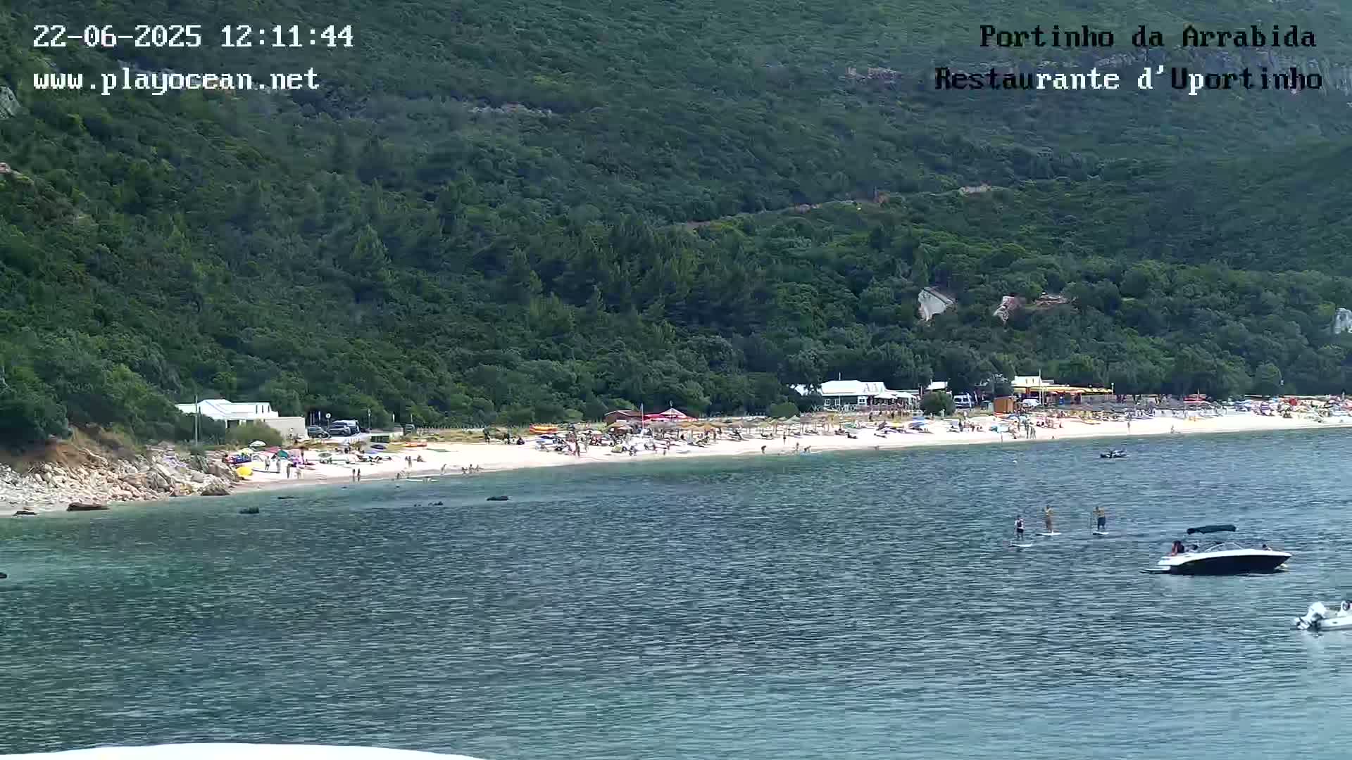 A sandy beach with people sunbathing and swimming is backed by lush green hills, and several small boats are visible on the calm water.