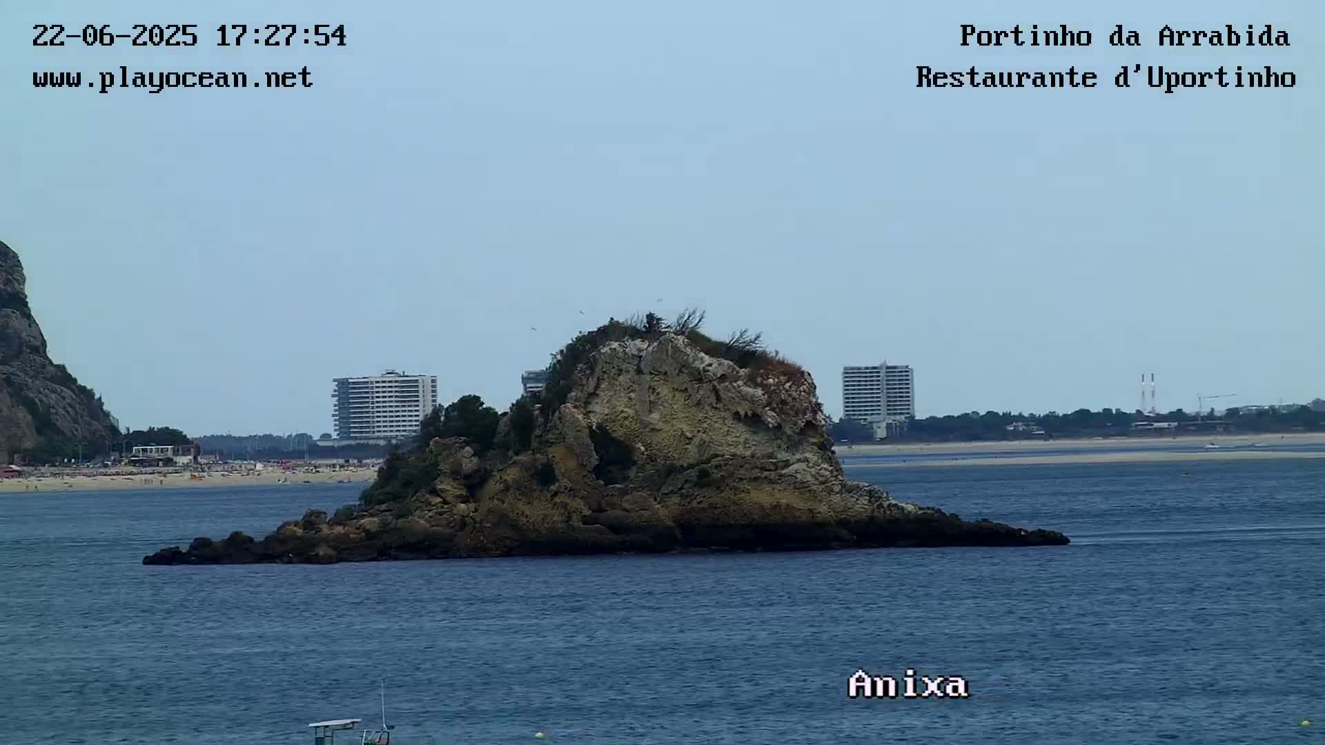 A rocky islet sits in calm blue water, with a sandy beach and buildings visible in the background under a clear sky.