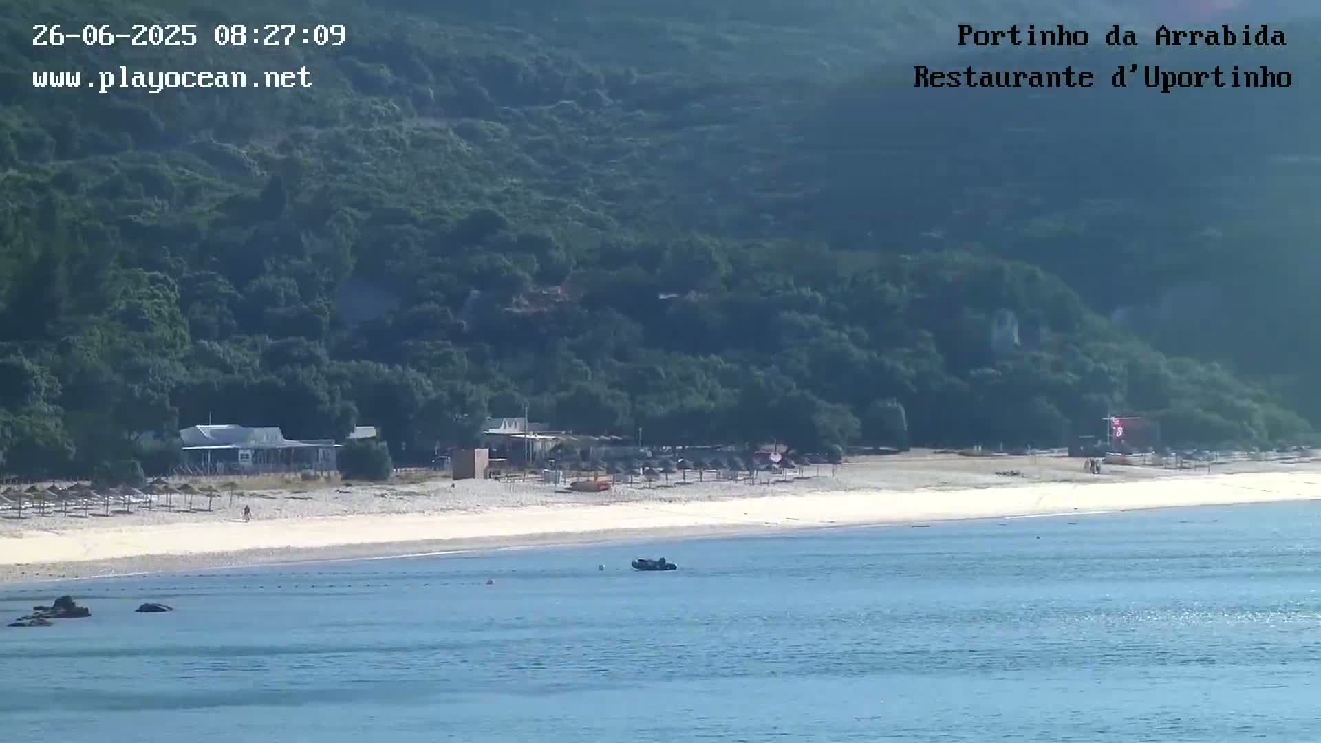 A sandy beach with calm, blue water, a few small buildings, and a green hillside in the background under sunny skies.