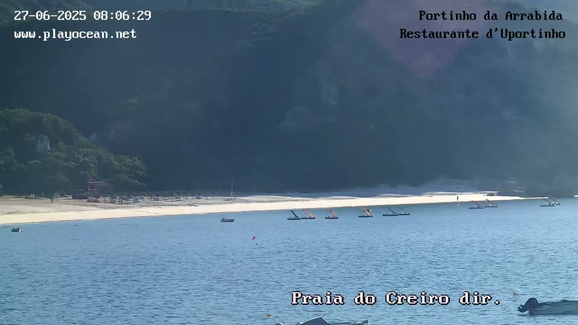 A sandy beach with several small boats in calm water is nestled below green hills on a sunny day.