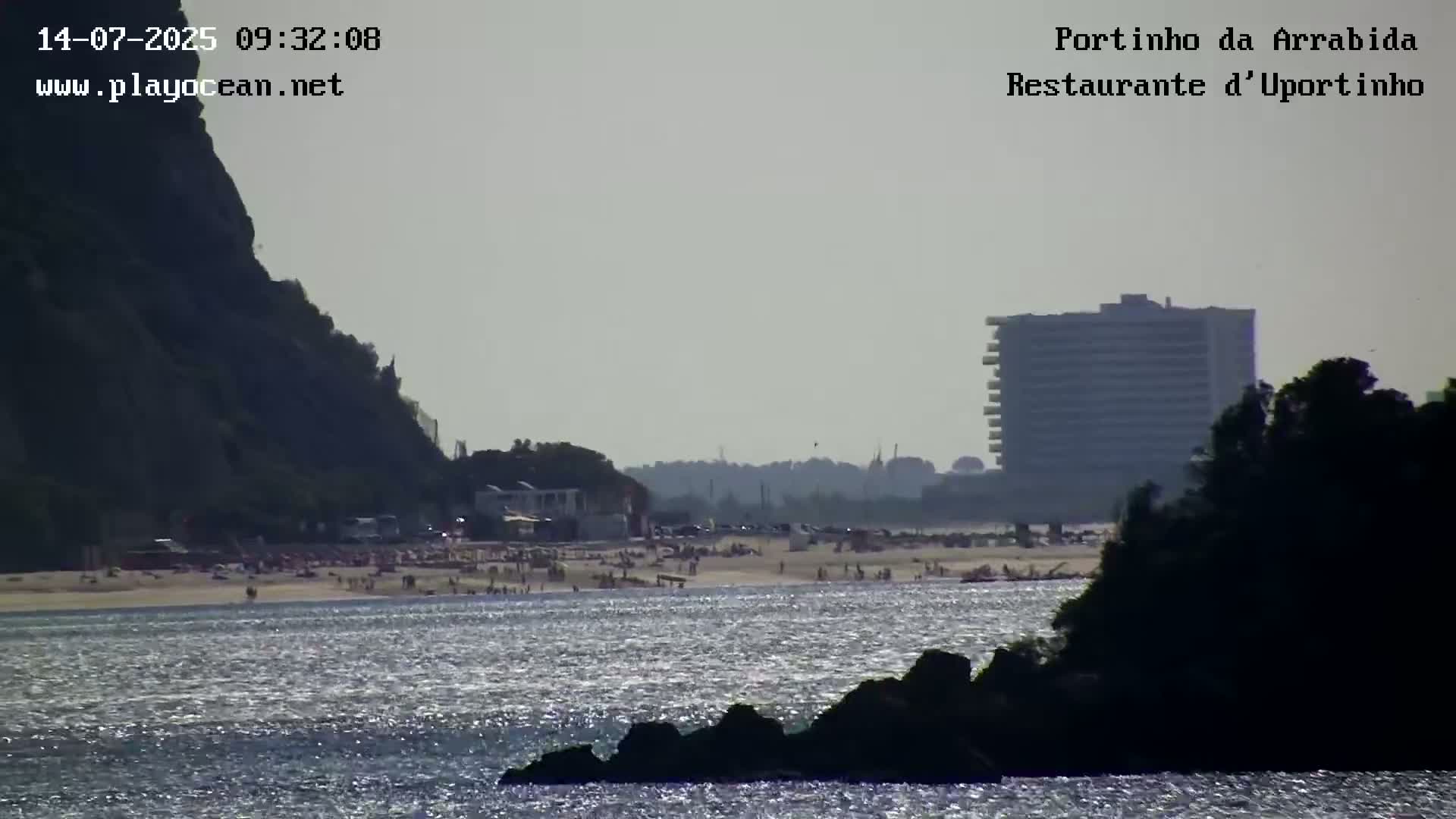 A hazy, sunny day reveals a beach crowded with people, a large building in the distance, and dark, rocky formations in the foreground of a calm body of water.