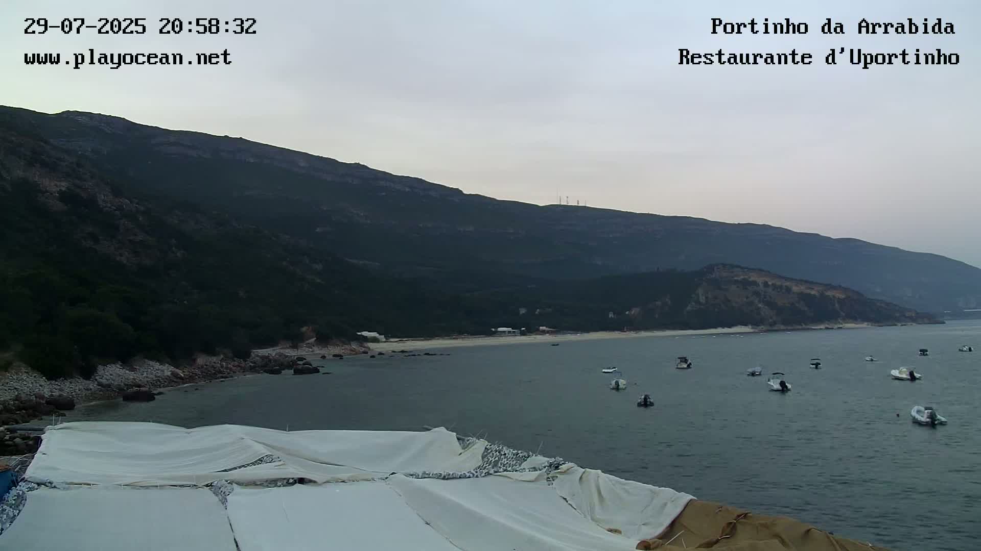 A partly cloudy day reveals a calm bay with several small boats, nestled against a verdant mountainside and a sandy beach.