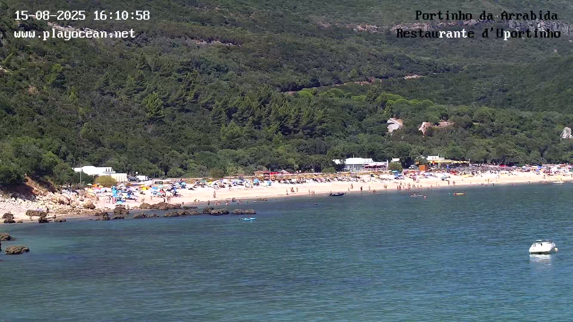 A partly cloudy day reveals a calm bay with several small boats, nestled against a verdant mountainside and a sandy beach.