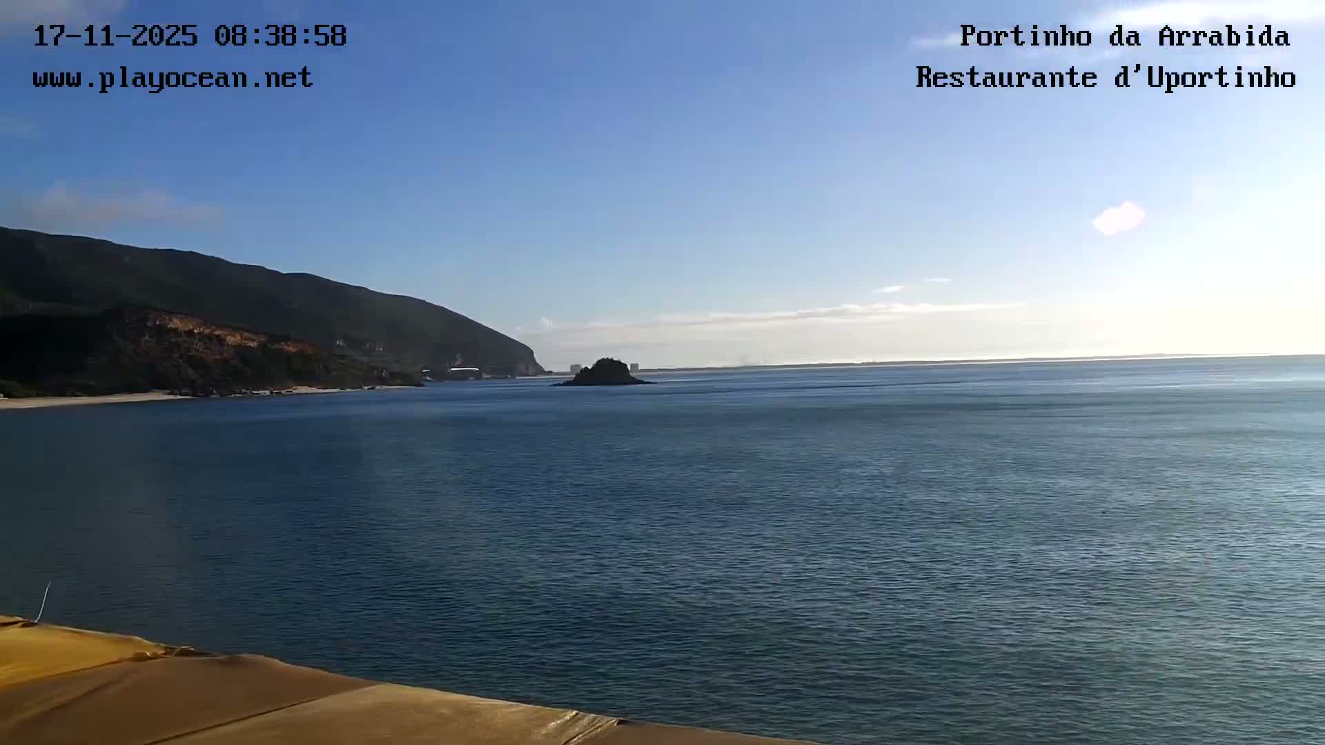 On a clear and sunny day, a calm blue ocean extends to the horizon, bordered on the left by a green, mountainous coastline with sandy patches and featuring a small dark rocky island in the mid-distance under a bright blue sky with a few wispy clouds.