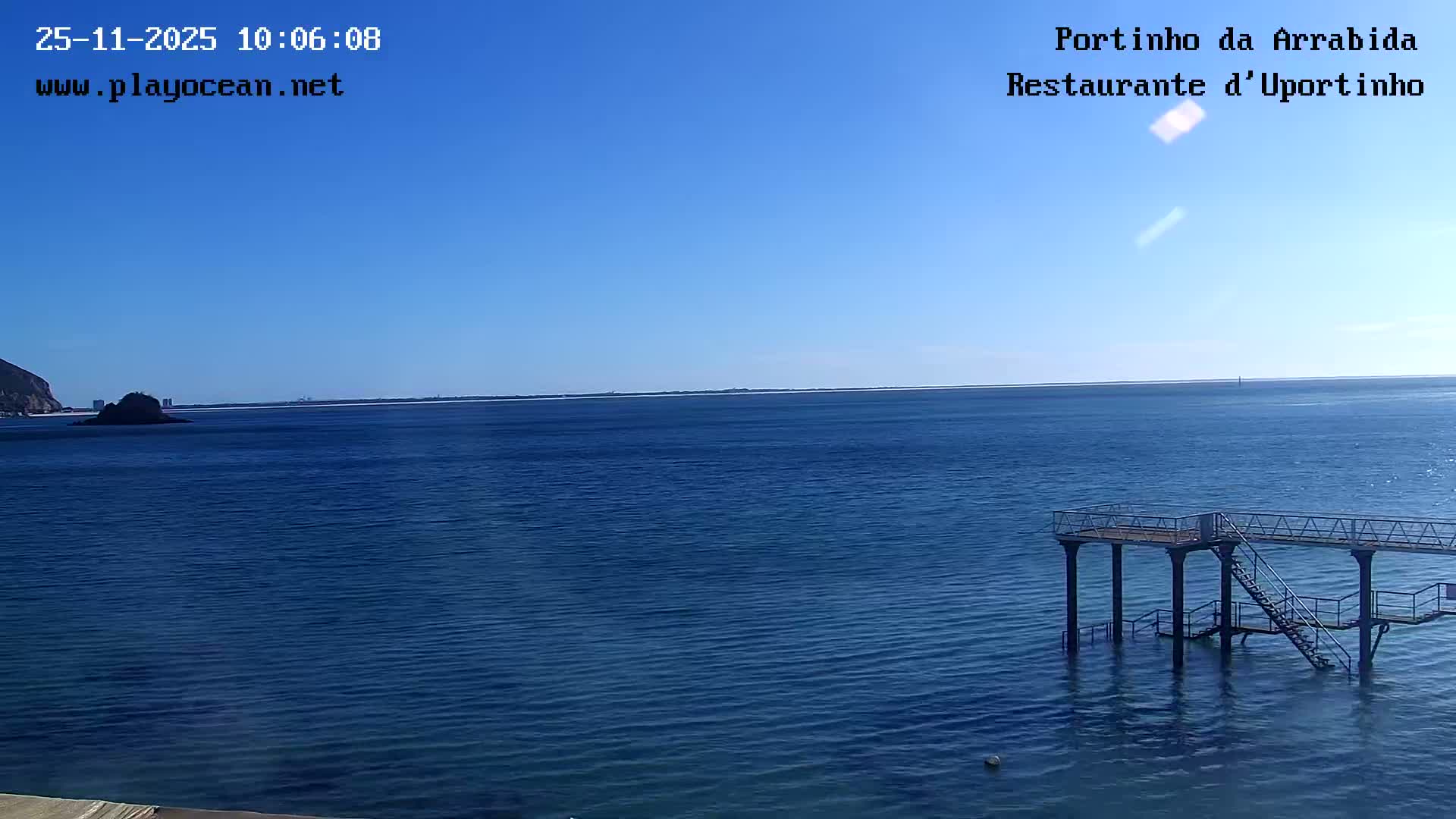 A clear blue sky extends over a tranquil blue ocean, where a multi-level metal platform with stairs descends into the water on the right, and a mountainous coastline is visible in the distance on the left.