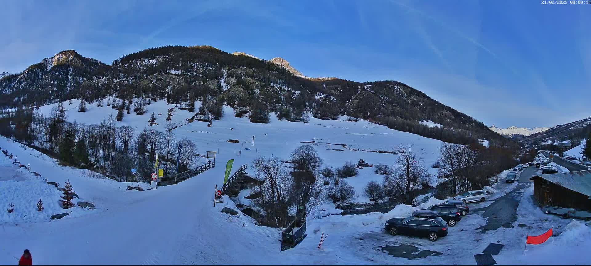 A snow-covered mountain slope with a few cars parked near a small building, under a clear blue sky.