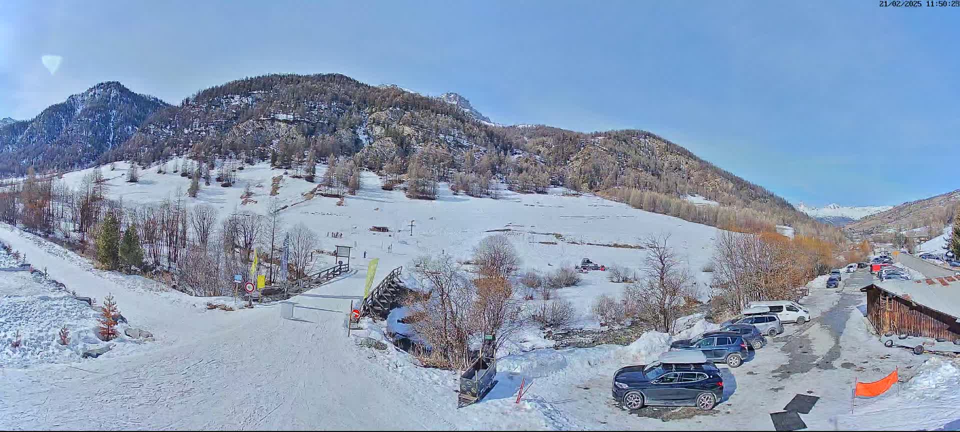 A snow-covered mountain valley on a sunny day, with a parking lot containing several cars near a wooden building.