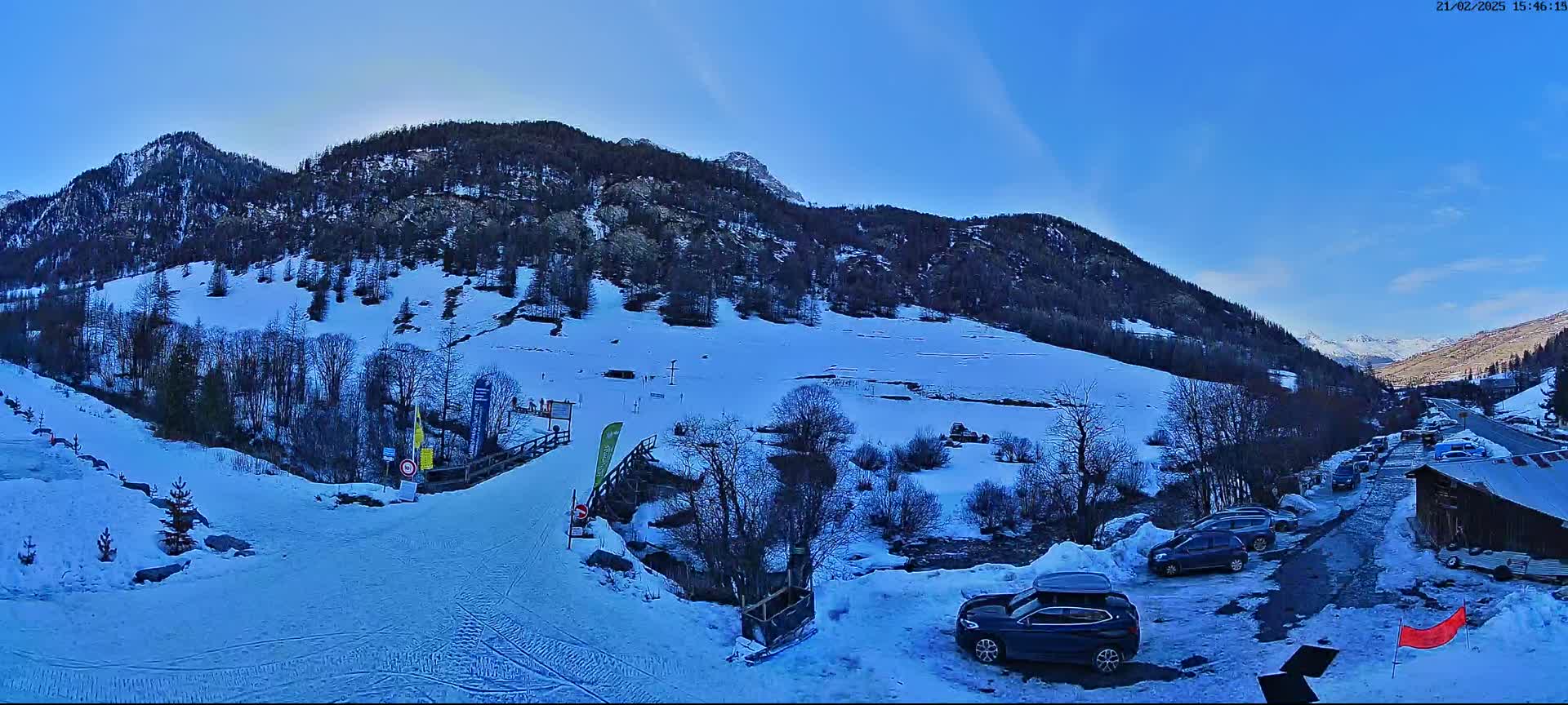A snow-covered mountainside with a ski slope and several cars parked in a lot under a clear blue sky.