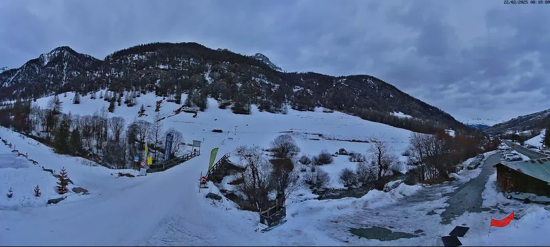 A snow-covered mountain valley under a cloudy sky shows a road leading to a small building and a ski slope.