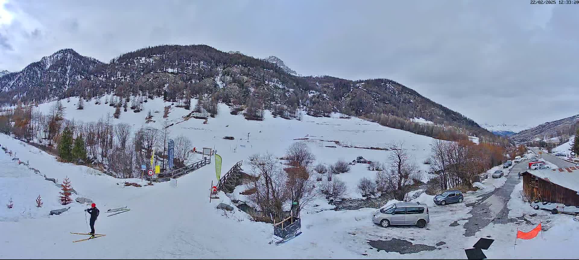 A snowy mountain valley scene with a few cars parked near a wooden building, a cross-country skier on a groomed track, and overcast skies.