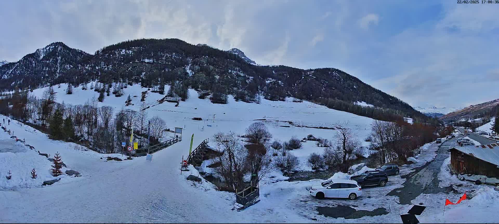 A snowy mountainside with a small parking lot and several cars is visible under a partly cloudy sky.