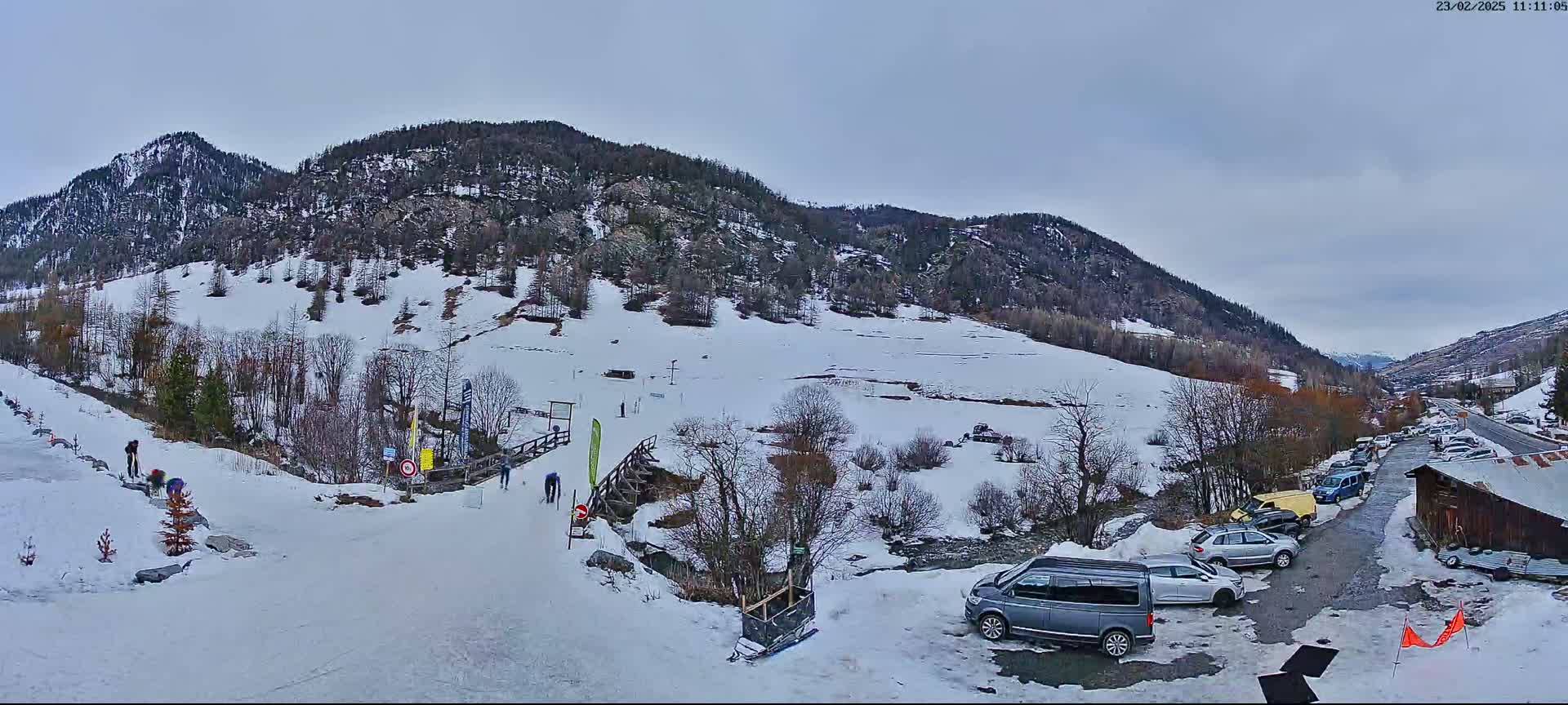 A snow-covered mountain valley scene on an overcast day shows a small parking lot with several cars and vans near a wooden building, with a few people visible on a snow-covered path leading up a hillside.