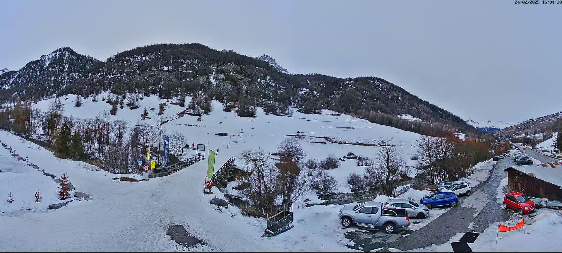 A snow-covered mountain valley scene with several vehicles parked near a wooden building under an overcast sky.