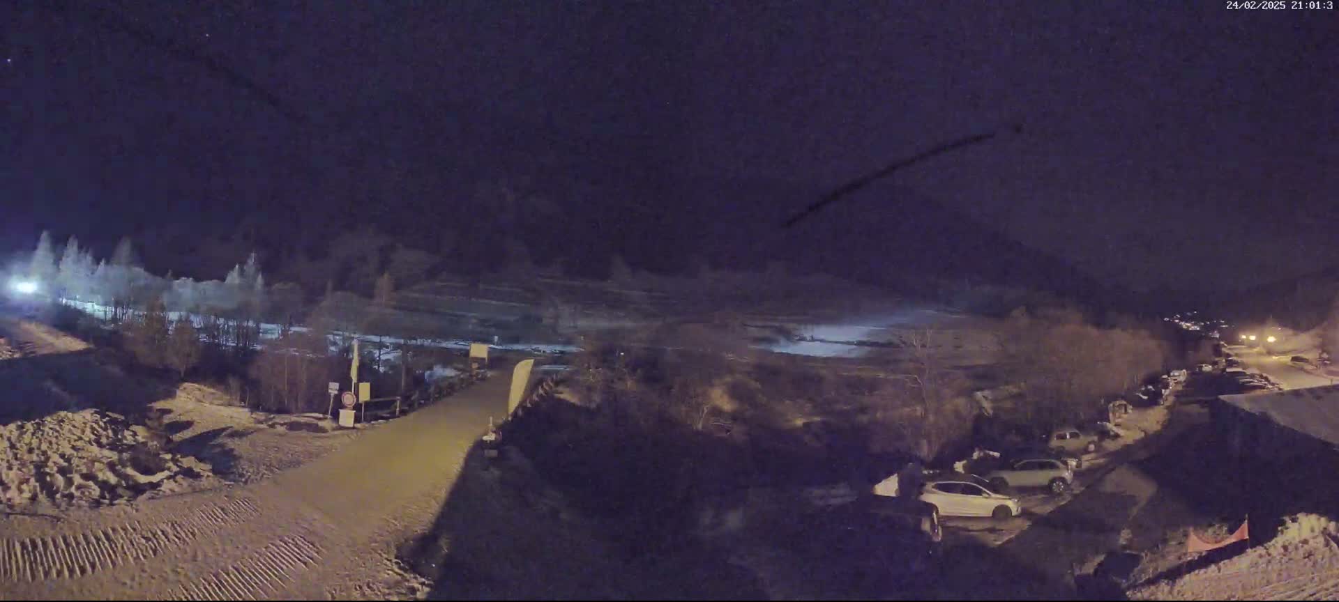 A nighttime, snow-covered parking lot and road are visible, with a few cars parked and a dark, snow-covered landscape in the background under a clear night sky.