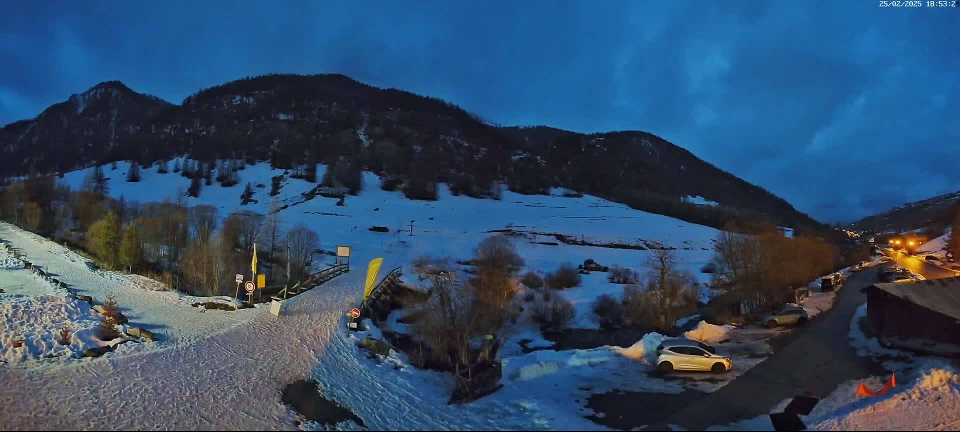 A snow-covered mountainside at dusk, with a small parking lot and a road leading to a wooden structure.