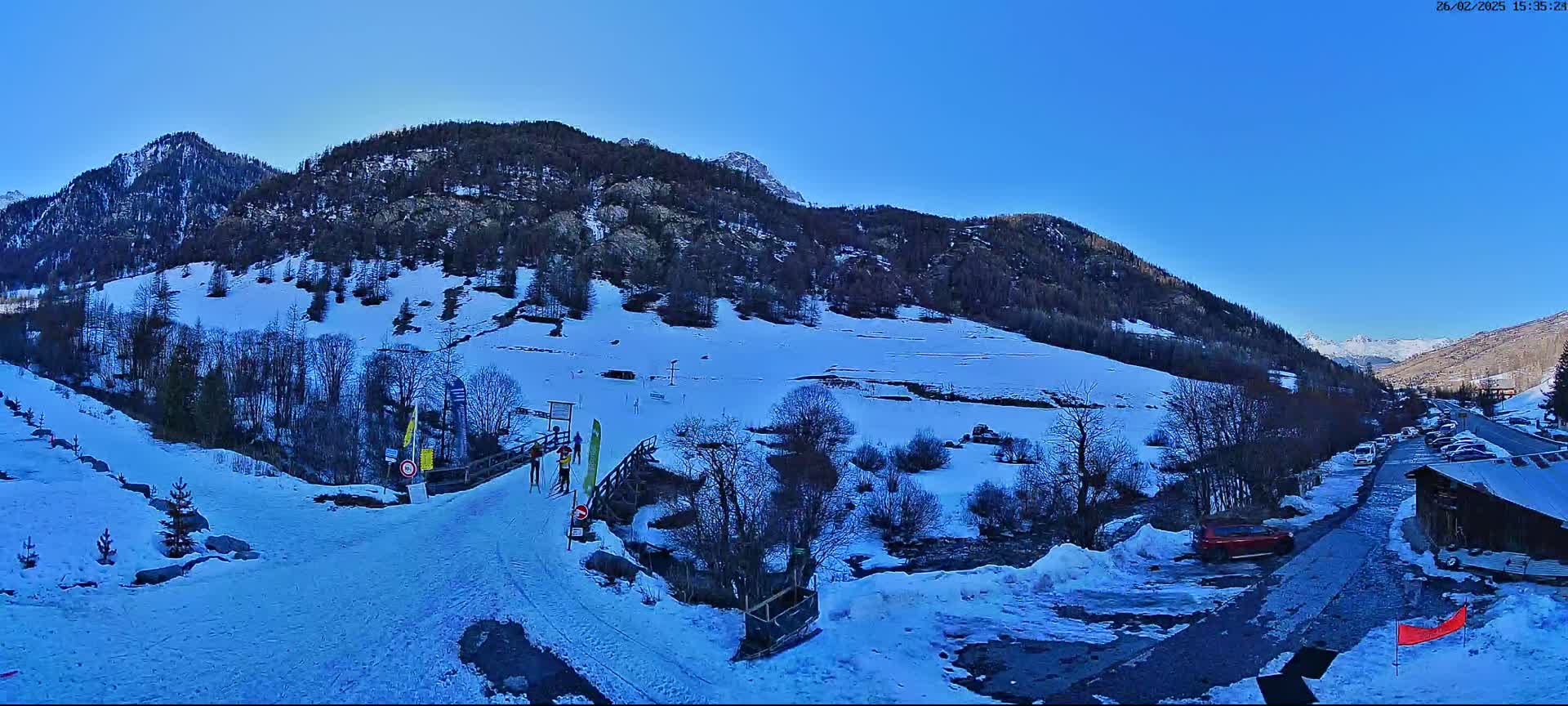 A snowy mountainside with a few cross-country skiers on a trail near a parking lot under a clear blue sky.