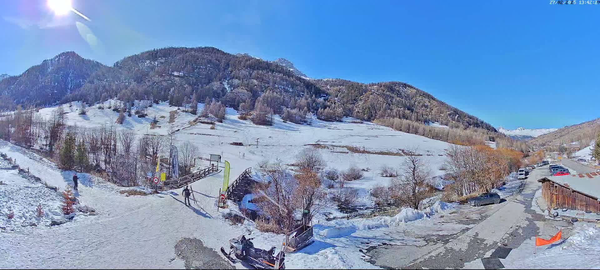 A sunny day in a snow-covered mountainous region shows a small village with parked cars near a snow-covered path leading to a ski slope.