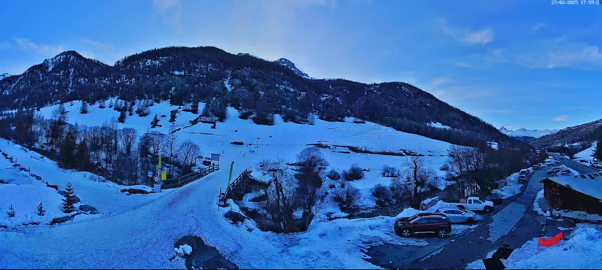 A snow-covered mountainside with a few cars parked near a small building under a clear blue sky.