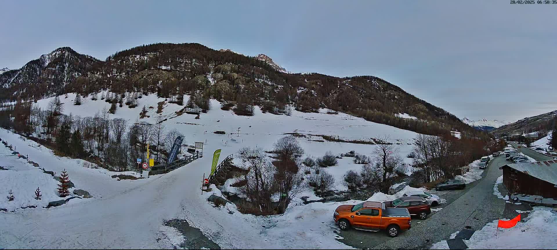 A snow-covered mountain valley scene under a pale sky shows a parking lot with several vehicles near a roadway leading to a snow-covered path and a wooden bridge.