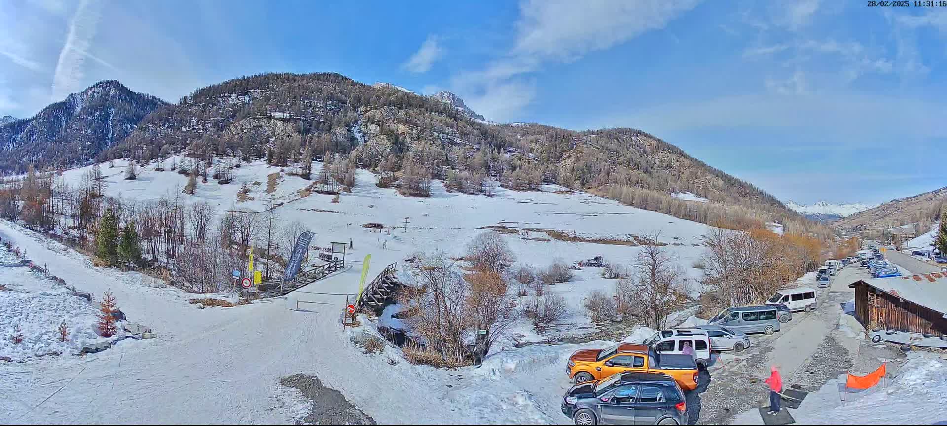 A snow-covered mountainside with a small parking lot of cars and a ski lift in view on a sunny day.