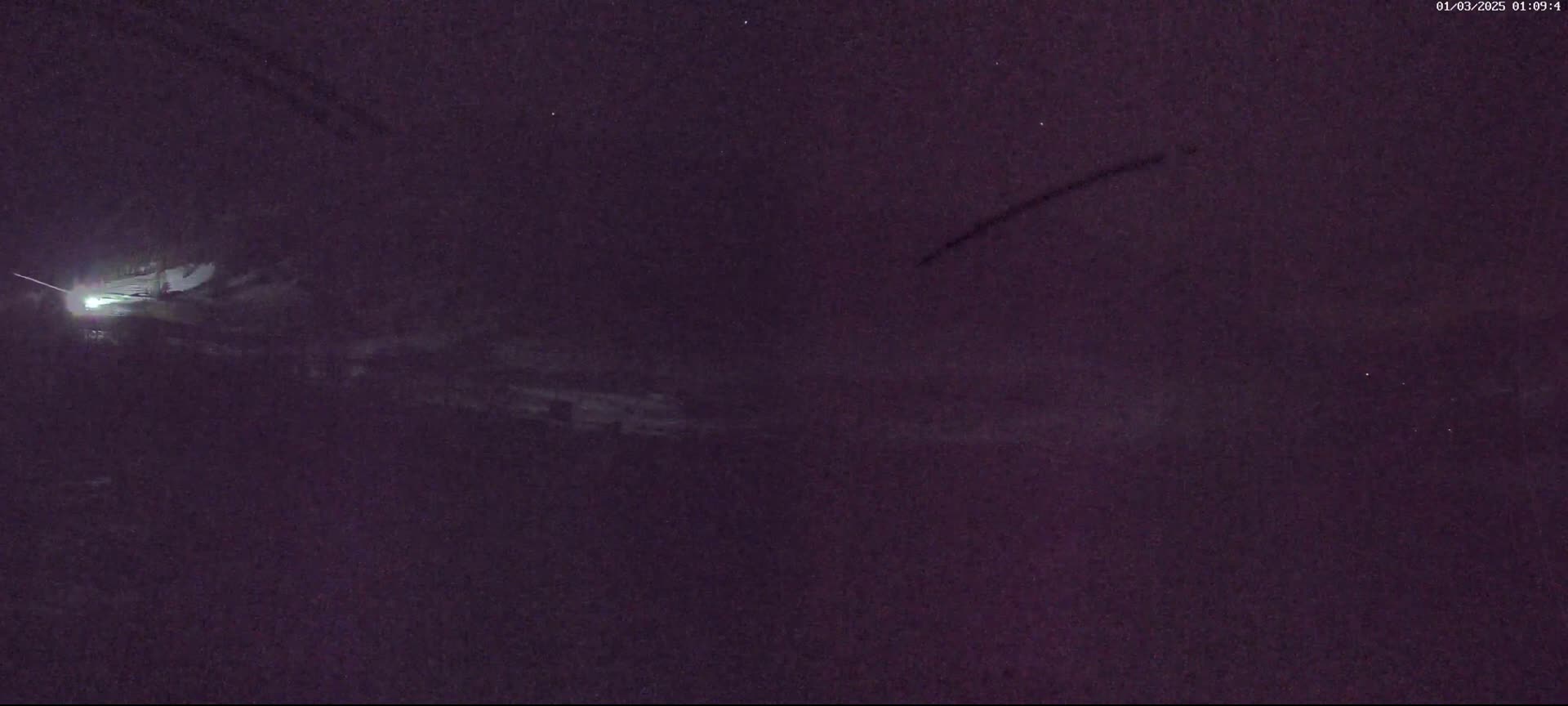 A nighttime, low-light view shows a faint, bright light source in the distance, possibly a vehicle's headlights, and dark, possibly snowy or icy, ground in front of it.