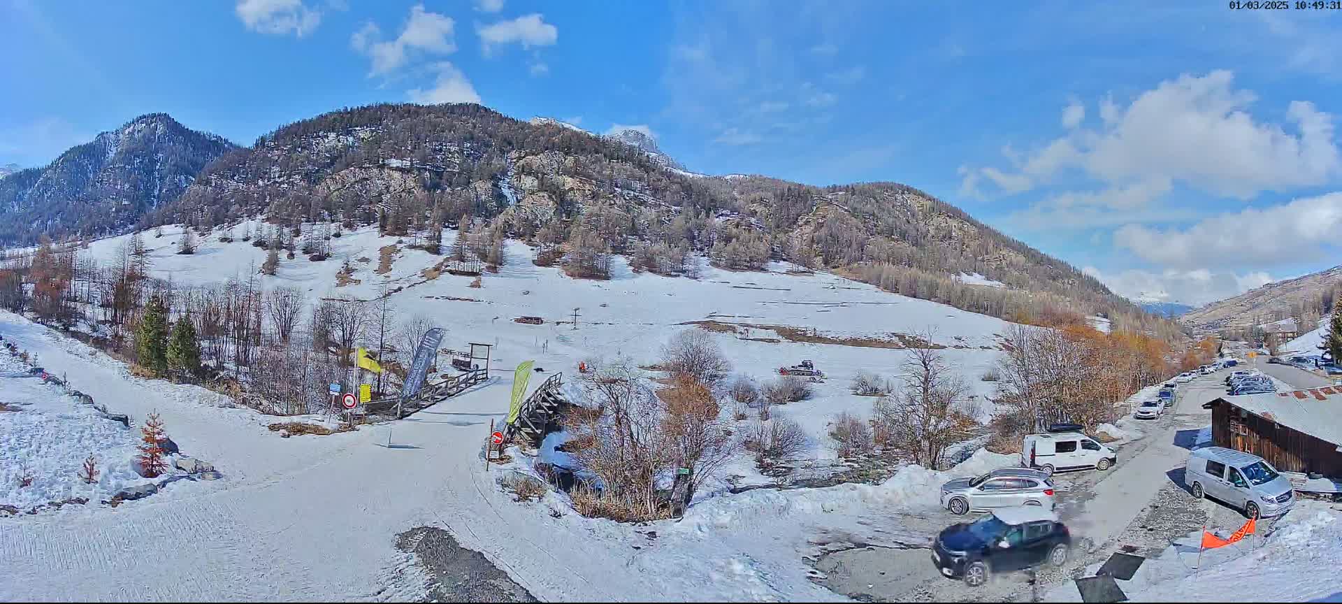 A snowy mountain valley scene on a partly sunny day shows a small ski area with a parking lot containing several cars and vans.