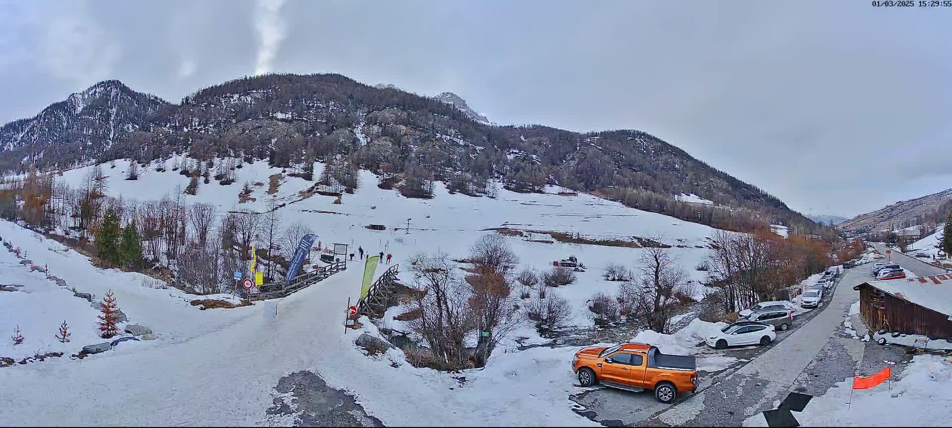 A snow-covered mountainside with a parking lot, a few cars, and an orange pickup truck, under an overcast sky.