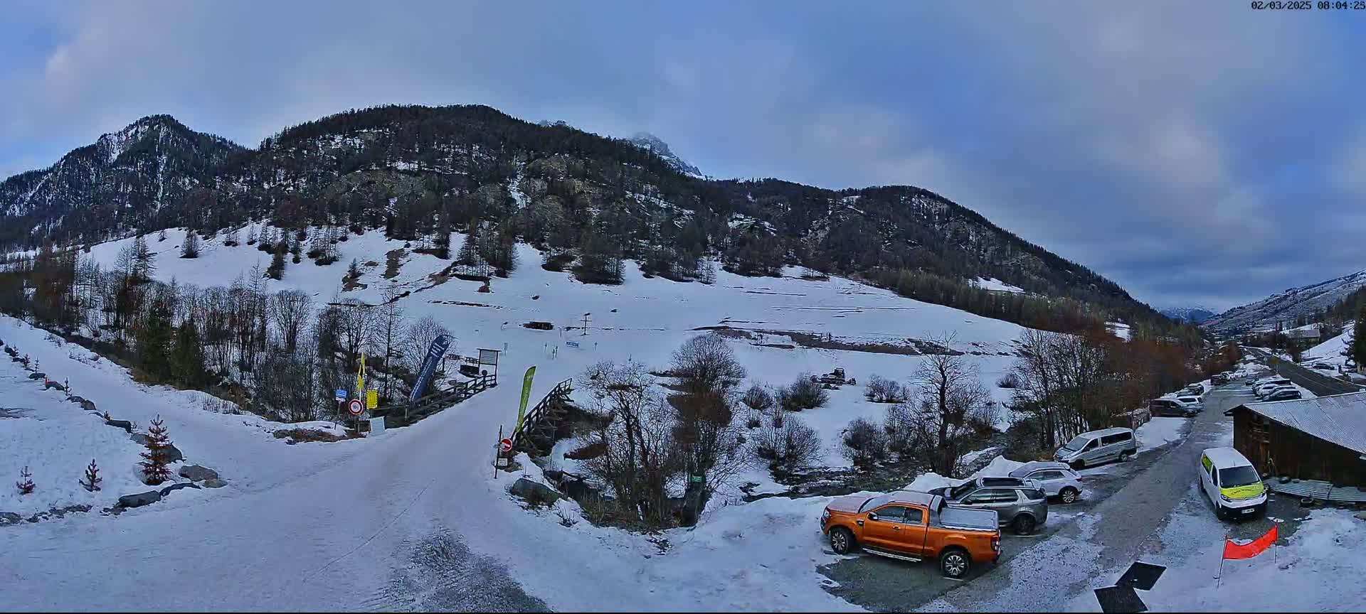 A snow-covered mountain valley scene with several vehicles parked near a small wooden building under a mostly cloudy sky.