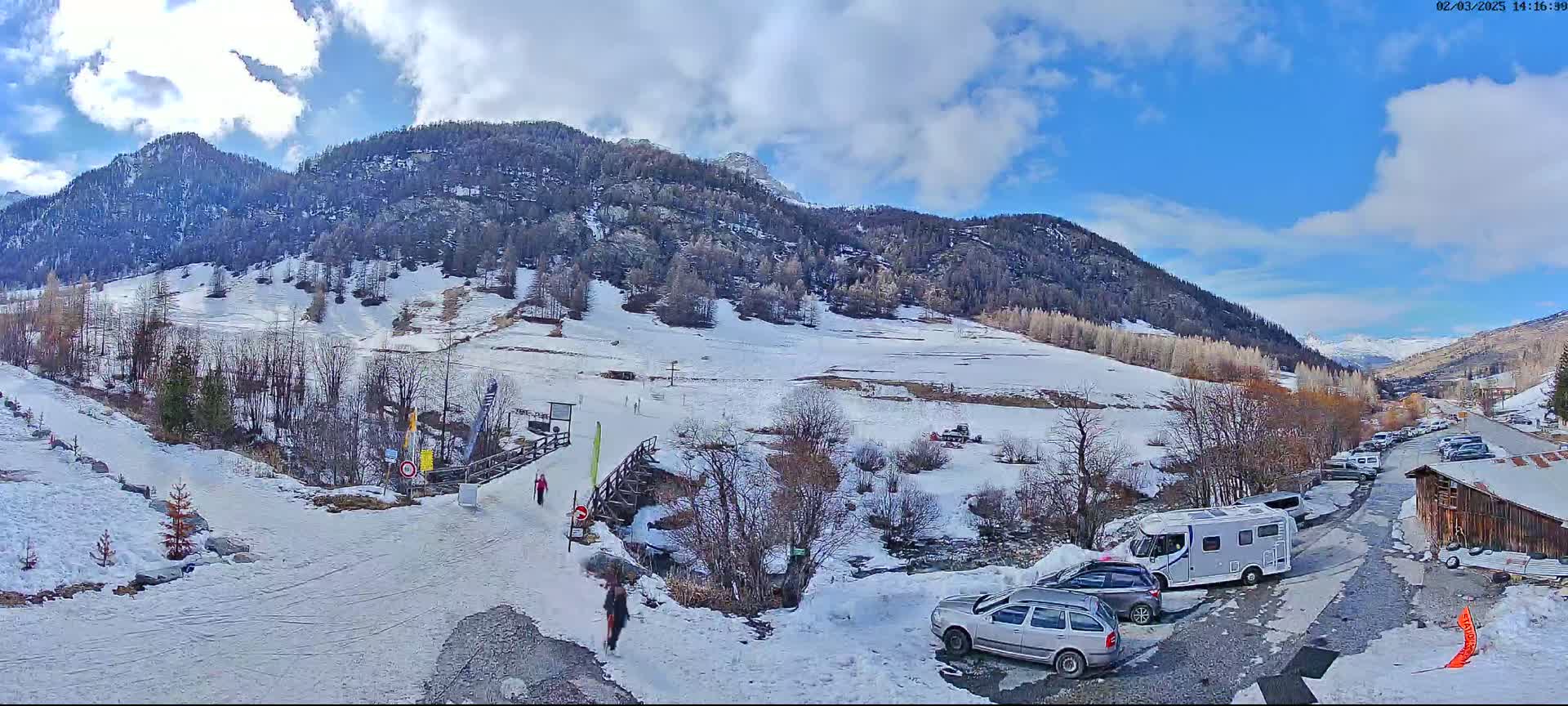 A snow-covered mountain valley scene on a partly sunny day, with a parking lot containing several cars and a camper van near a wooden building.