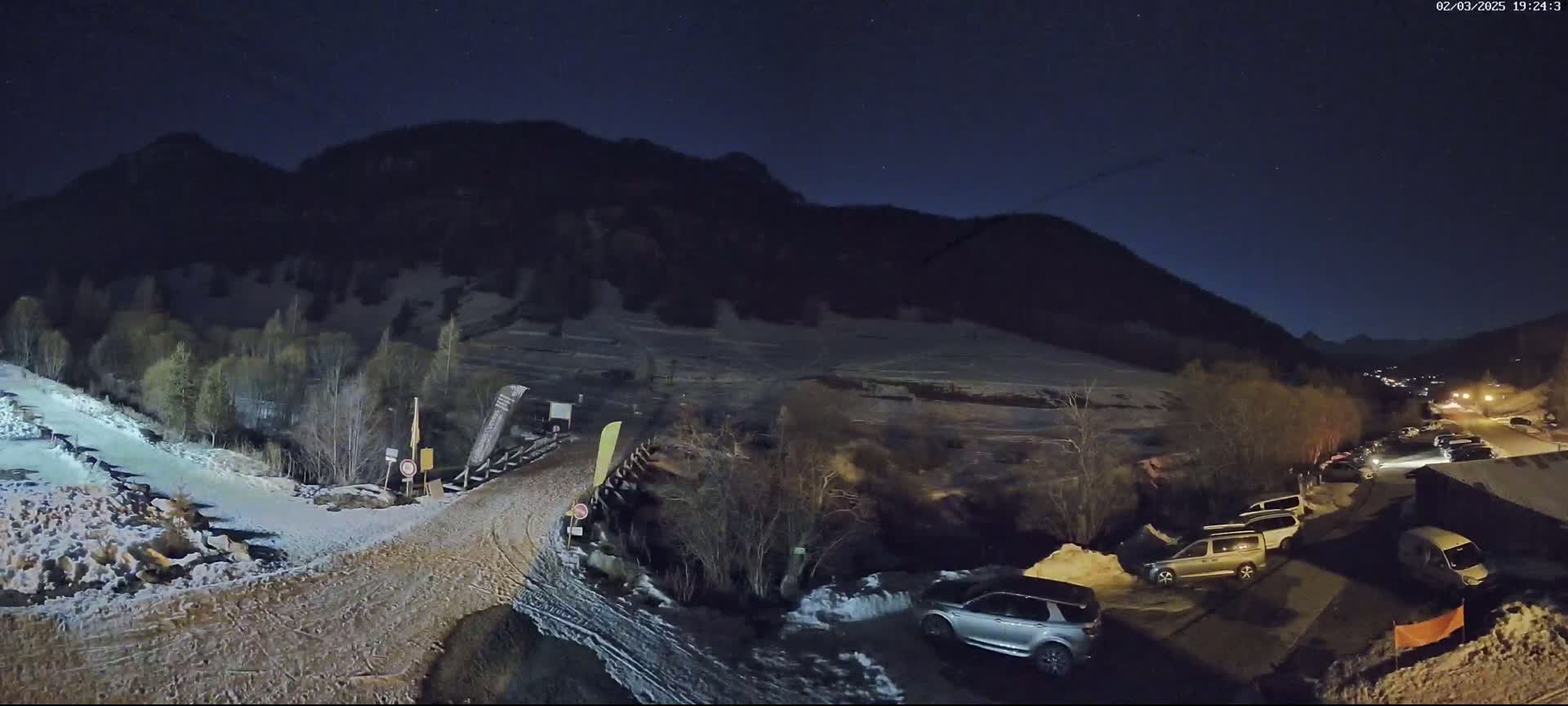 A nighttime scene shows a snow-covered parking lot with several vehicles near a mountain under a clear, starry sky.