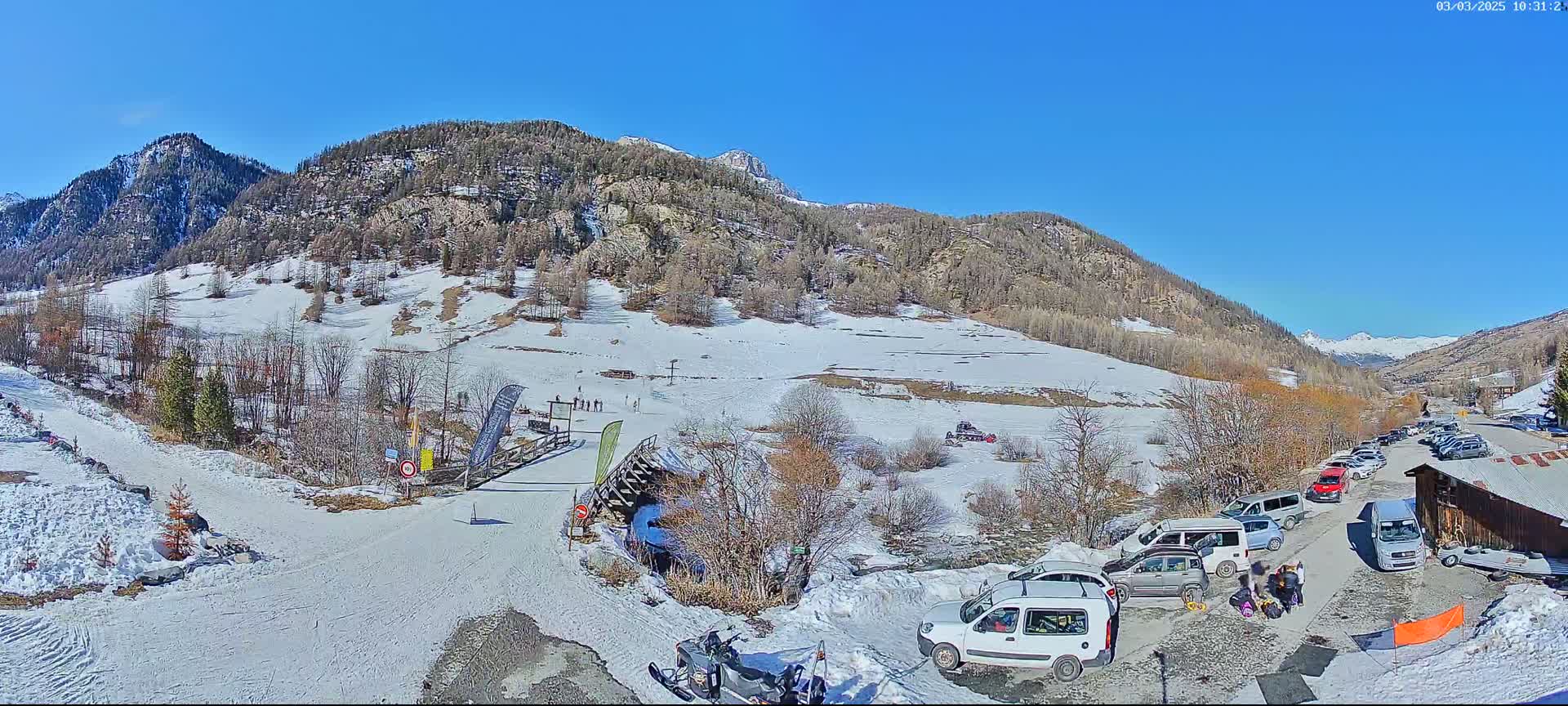 A snow-covered mountain valley scene on a sunny day, with cars parked near a small building and a snowmobile visible in the foreground.