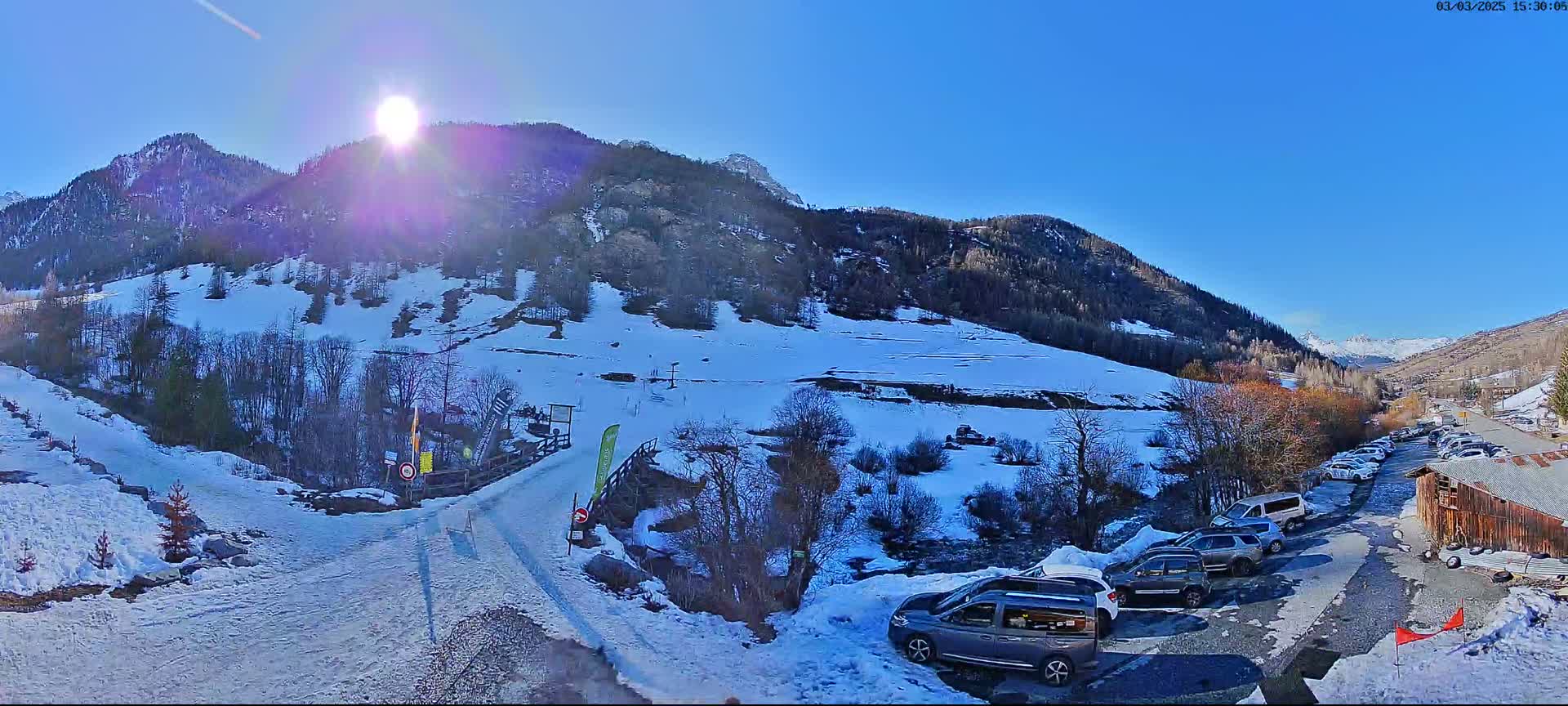 A sunny day in a snow-covered mountain valley shows a parking lot with several vehicles near a rustic building, with a snow-covered slope and ski lift in the background.